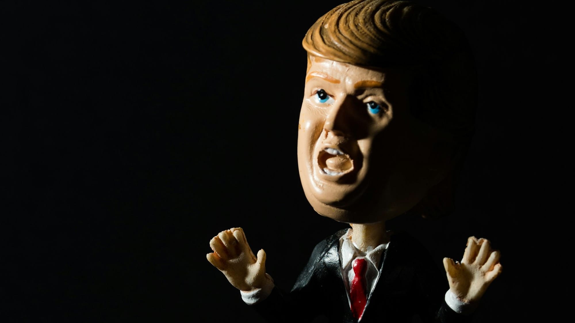 A bobblehead of Donald Trump, hands gesturing and mouth open, set against a black background.